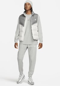 Inimene, kellel on peal hall nokamüts, kahes toonides hall ja valge sulevest, hall pusa, sobivad trenni püksid ja valged Nike tennised.