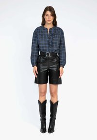 Chemise à carreaux bleus à manches longues avec volants, shorts noirs en cuir taille haute, et bottes de cowboy noires hautes avec des détails en relief.