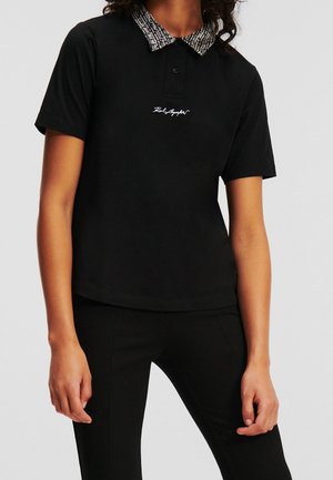 Polo noir à manches courtes, avec un col à motif texturé et un logo brodé sur la poitrine. Le tissu semble lisse et ajusté.