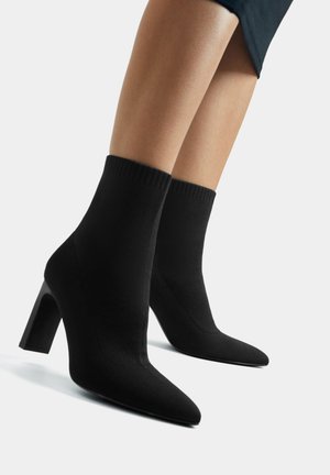 Nogi w czarnych dzianinowych botkach typu sock boots z szpiczastymi noskami i blokowymi obcasami, na białym tle.