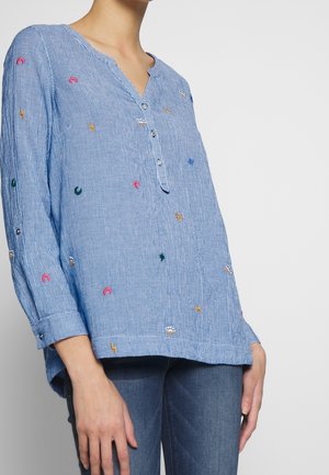 Frau trägt ein blau kariertes Langarmhemd mit kleinen bunten Stickereien und drei Knöpfen am Ausschnitt, kombiniert mit blauen Jeans.