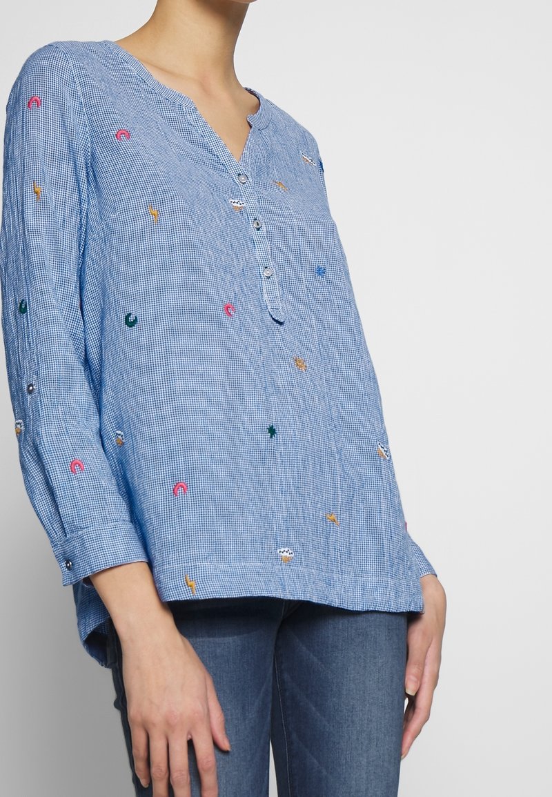 Femme portant une chemise à manches longues à carreaux bleus avec de petites formes brodées colorées et trois boutons au niveau de l'encolure, associée à un jean bleu.