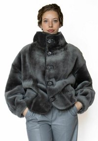 Grijze nepbontjas met een hoge kraag, voorzien van grote knopen, een afgerond ontwerp en volumineuze mouwen. Gecombineerd met grijze leren broek.