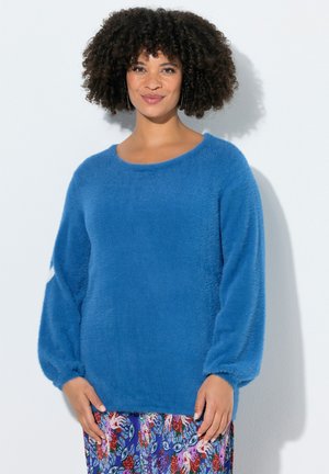 Kvinde med krøllet hår iført en blå, blød, langærmet sweater og en farverig blomsterprintet nederdel, stående mod en ensfarvet hvid baggrund.