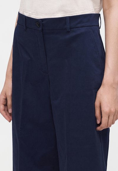 Personne portant un pantalon taille haute bleu marine avec poches latérales et fermeture boutonnée à l'avant, associé à un haut blanc.