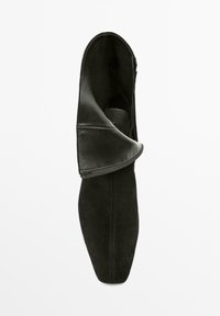 Stivale in camoscio nero con patta a risvolto in pelle e dettagli con cuciture in rilievo, visto dall'alto su uno sfondo bianco.