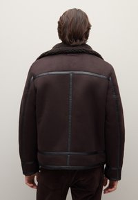 Braune Lammfelljacke mit schwarzen Akzenten, ausgestattet mit einem breiten Kragen und einem geometrischen Paneeldesign. Weiche Textur mit auffälligen Stickdetails.