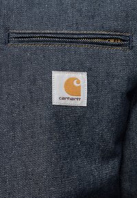 Σκούρο μπλε υφαντό τζιν με οριζόντια τσέπη με φερμουάρ και λευκή ετικέτα με κίτρινο λογότυπο. Ραφές με αντίθετα χρώματα.