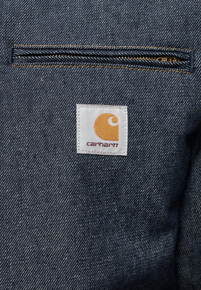 Tissu denim bleu foncé avec une poche à fermeture éclair horizontale et une étiquette blanche comportant un logo jaune. Accents de couture en couleurs contrastantes.