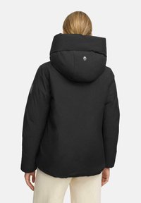 Vrouw die een zwarte pufferjas met een grote capuchon draagt, gezien van achteren, in combinatie met lichtgekleurde broeken.