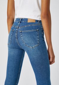 Los jeans skinny de mezclilla azul presentan una cintura de media altura, dos bolsillos traseros y una etiqueta de marca de cuero marrón en la cintura. Textura suave y elástica.