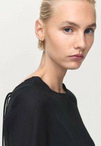 Mujer rubia con ojos azules y pequeños pendientes de aro dorados lleva una blusa negra, mirando ligeramente hacia un lado con una expresión neutral.