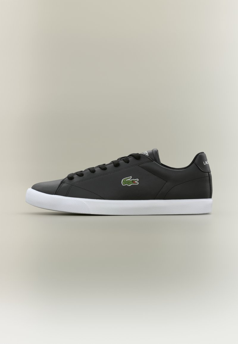 Baskets basses noires avec semelle blanche, lacets à l’avant et logo crocodile vert sur le côté, présentées de profil sur un fond neutre.
