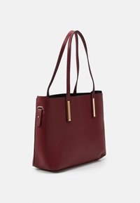 Bordeaux kožená kabelka s dlouhými ušima a zlatými obdélníkovými akcenty na předních popruzích, zobrazená na bílém pozadí.