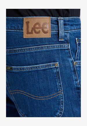 Közeli kép a hátsó zsebről és a derékrészről kék farmereken, melyen barna bőr Lee márkajelzés található a zseb felett varrva.