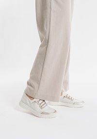 Weiße Sneakers mit strukturiertem Obermaterial, beigen Akzenten und flachen Sohlen. Kombiniert mit hellbeigen weiten Hosen.