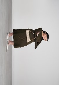 Kobieta w ciemnooliwkowej midi sukience i białych szpilkach, trzymająca małą beżową torbę na łańcuszku na jednolitym tle.