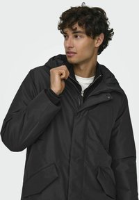 Chaqueta negra con capucha, cierre frontal de cremallera, grandes bolsillos frontales y textura suave. Presenta un diseño en capas y costuras visibles.