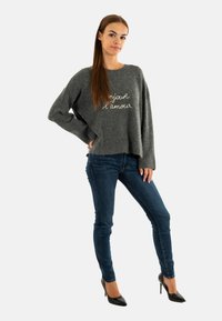 Grijze gebreide trui met de tekst "bonjour l'amour", gecombineerd met donkerblauwe skinny jeans en zwarte hoge hakken. Zachte stof en een ontspannen pasvorm.