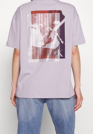 Osoba nosząca liliową koszulkę z prostokątnym nadrukiem twarzy, na którym powtarza się tekst "Calvin Klein Jeans" na plecach, w połączeniu z niebieskimi jeansami.