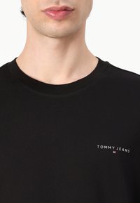 T-shirt en coton noir avec un col rond et un petit logo blanc "TOMMY JEANS" avec un accent rouge et bleu sur le côté gauche.