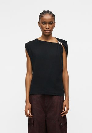 Γυναίκα με πλεξούδες που φοράει αμάνικο μαύρο τοπ με επενδεδυμένους ώμους και σκουρόχρωμο παντελόνι cargo, σε απλό φόντο.