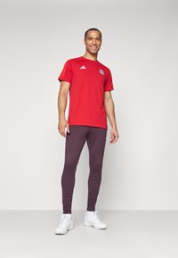Rød idretts t-skjorte med korte ermer og en logo, kombinert med tettsittende mørk lilla bukser og hvite joggesko. Enkel og ren design.