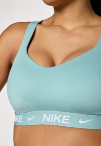 Svijetloplava sportska braletka s izrezom u obliku lopte, izrađena od glatkog materijala, s bijelom elastičnom trakom na kojoj je ispisano "NIKE".