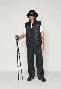 Crna podstavljena prsluk preko bijele majice, crne kombinezone i crne cipele. Crna kapa i sunčane naočale upotpunjuju izgled spreman za vani, držeći planinarske štapove.