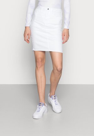 Kvinde iført en hvid denim nederdel, hvid langærmet top og hvide sneakers med lavendel accenter, stående på en grå gulv.