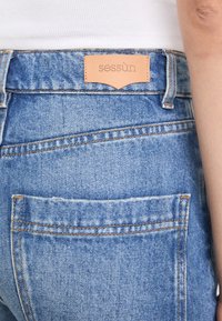 Vaqueros de mezclilla azul con una etiqueta de cuero que muestra la marca "sessùn" sobre un bolsillo trasero y detalles de costura visibles.