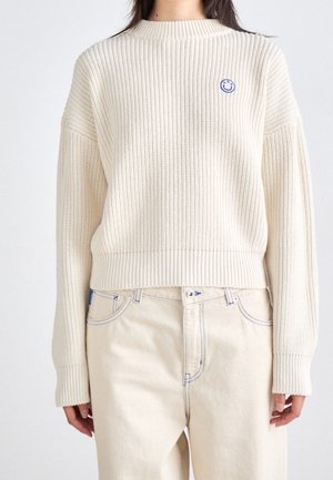 Crème, geribbelde trui met een ronde hals en verlaagde schouders. Voorzien van een klein blauw glimlach-patch op de borst. Gecast in combinatie met lichte broek.