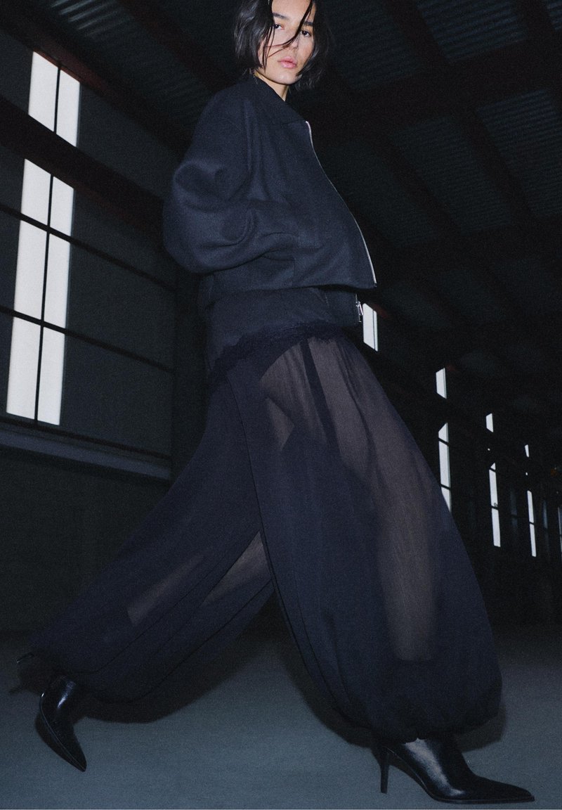 Jachetă bomber neagră cu mâneci largi, purtată peste o fustă neagră transparentă. Ținuta are un design fluid și este completată de cizme negre cu toc înalt.