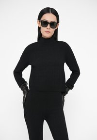 Pulover negru tricotat cu guler înalt, prezentând un model texturat în formă de diamant. Pantaloni negri mulați și mănuși din piele completează ținuta.