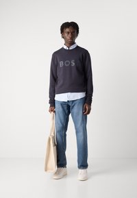 Ναυτικό φούτερ με έντονο λογότυπο "BOSS", σε στρώσεις πάνω από ένα ανοιχτό μπλε πουκάμισο με γιακά. Φοριέται με τζιν και κρεμ sneakers, κρατώντας μια μπεζ τσάντα.