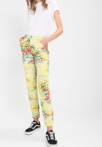 Blommönstrade, ljusgula byxor med gröna och rosa accenter, med sidofickor och tapered fit, kombinerat med svarta sneakers.