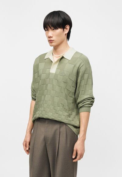 Νεαρός άνδρας που φοράει πράσινο καρό πλεκτό πουλόβερ με μπεζ γιακά, καφέ πλισέ παντελόνια και περιδέραιο με μαργαριτάρια, στέκεται μπροστά σε μονόχρωμο φόντο.