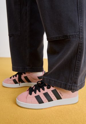 Personne portant des baskets Adidas Campus roses avec des bandes noires et des lacets noirs, debout sur une surface jaune avec un pantalon noir couvrant les chevilles.