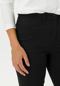 Pantaloni neri attillati con una texture liscia, dotati di una tasca frontale, chiusura con bottone e una piega discreta. Mano appoggiata sulla vita.