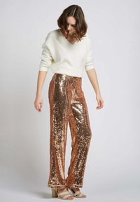 Pantalons en sequins rose gold avec une coupe ample, associés à un pull crème à col en V et des sandales à talons carrés beige clair. Texture lisse, finition brillante.