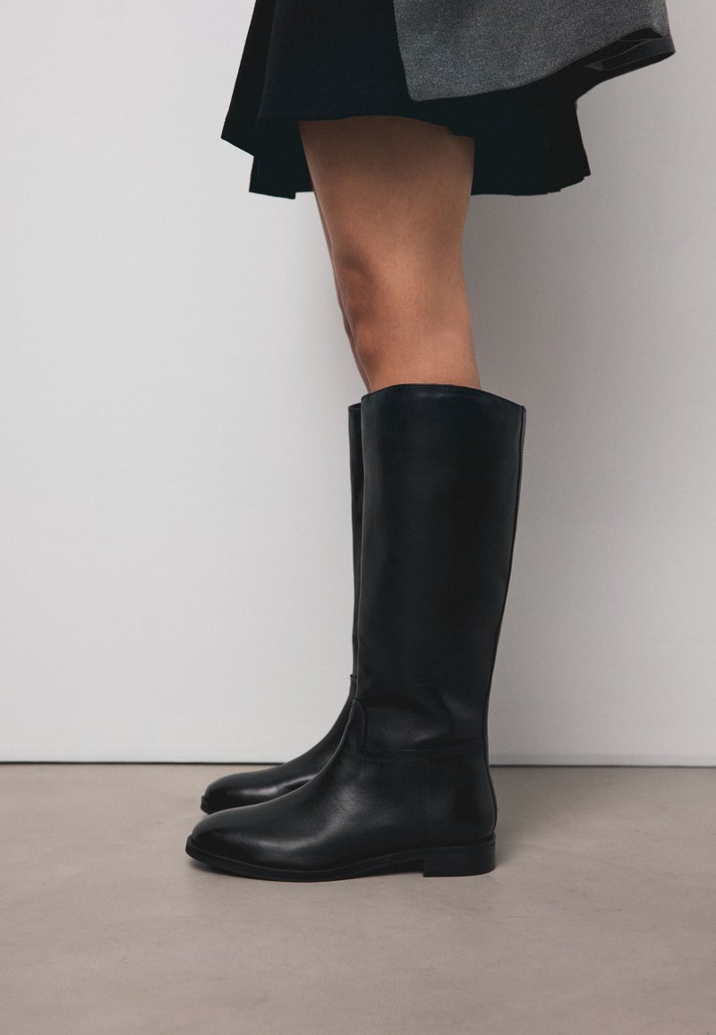 Osoba nosząca czarne skórzane kozaki do kolan i ciemną spódnicę, stojąca na gładkiej szarej podłodze na tle białej ściany.