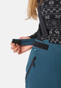 Läheltä kuvattu henkilö säätämässä sinisten ulkoiluhousujen tarranauhakaulusta, joissa on musta vetoketjupussi ja kuvioitu musta neule.