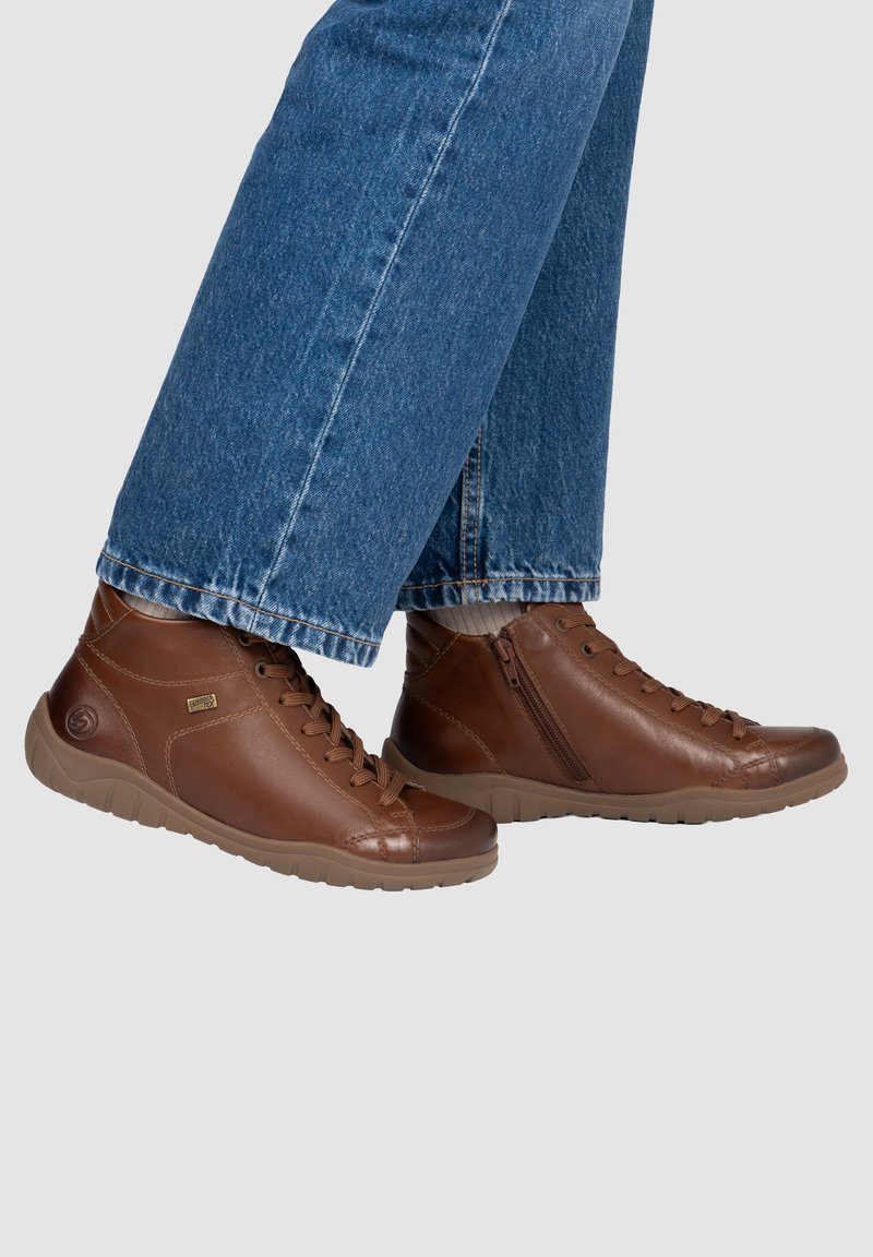 Bottines montantes en cuir marron à lacets avec fermeture éclair latérale, portées avec un jean en denim bleu et des chaussettes blanches sur fond gris clair.
