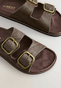 Sandales marron avec deux larges brides présentant des boucles carrées en laiton et une semelle intérieure souple sur un fond neutre.