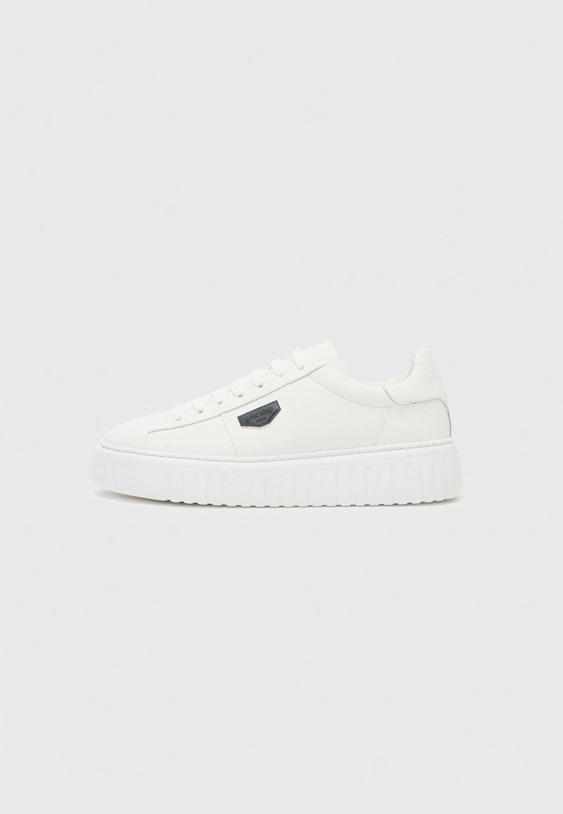 White sneakers with a smooth leather upper, flat lace closure, and a textured rubber sole featuring vertical grooves. Metal logo accent on side.