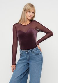 Een jumpsuit met een hartvormige fluwelen bodice in diep bordeaux, gecombineerd met lange doorschijnende mesh mouwen, gestopt in high-waisted blauwe spijkerbroek.