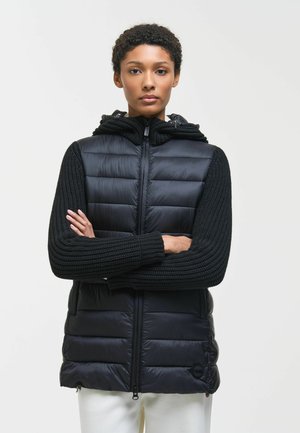 Donna con capelli corti e ricci che indossa un gilet imbottito nero con maniche e cappuccio lavorati a maglia, in piedi con le braccia incrociate davanti a uno sfondo bianco.