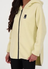 Ljusgult fleece-hoodie med texturerad yta, frontdragkedja och sidorevor. Har en liten svart logotyp på bröstområdet.