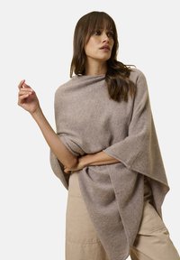 Donna con lunghi capelli castani che indossa un poncho beige largo e pantaloni beige, in posa con un braccio piegato e guardando di lato.