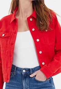 Veste en denim rouge avec quincaillerie argentée, poches avant et coupe ajustée. Portée sur un haut blanc et associée à un jean bleu taille haute.